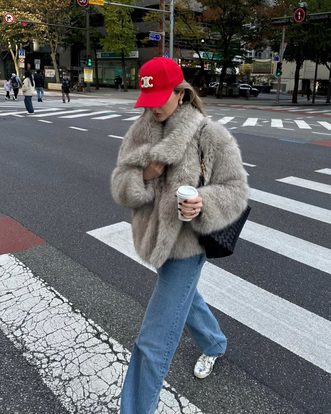 Person wearing a red cap and gray fur coat crossing a street.
