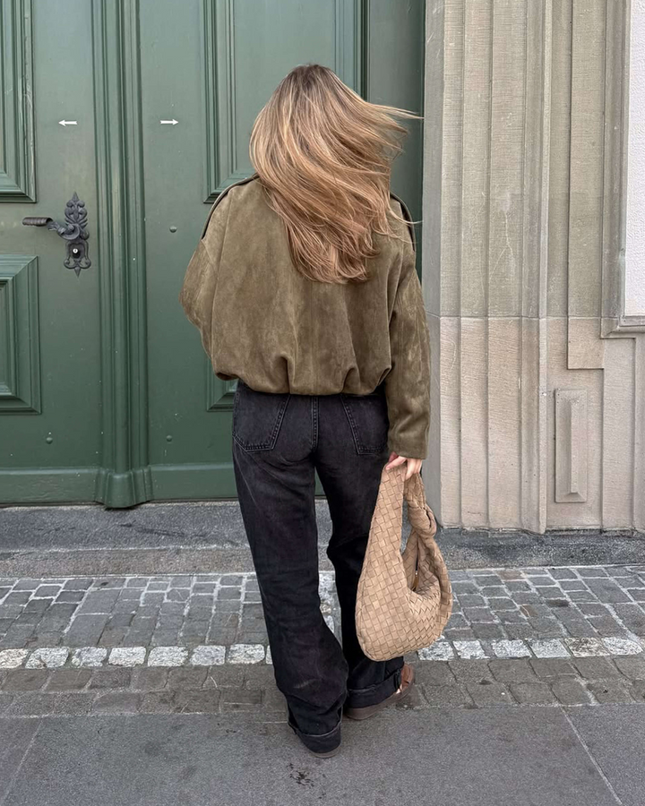 Person wearing a green jacket and dark pants, holding a beige bag, standing in front of a green door.