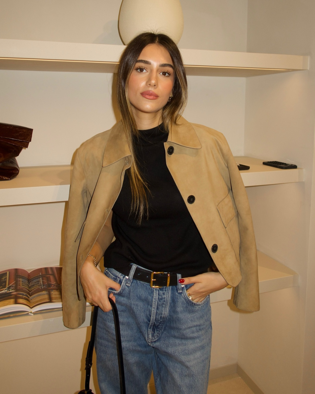 Woman wearing a beige coat, black top, and blue jeans indoors.