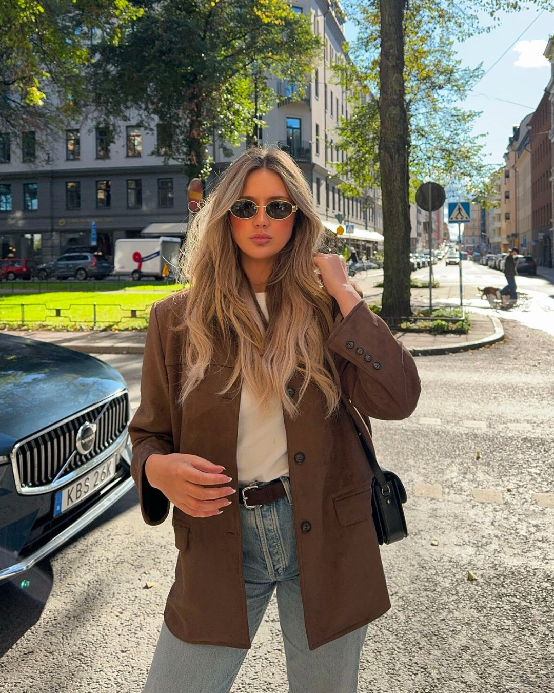 Woman in a brown coat standing on a city street with cars and buildings in the background.