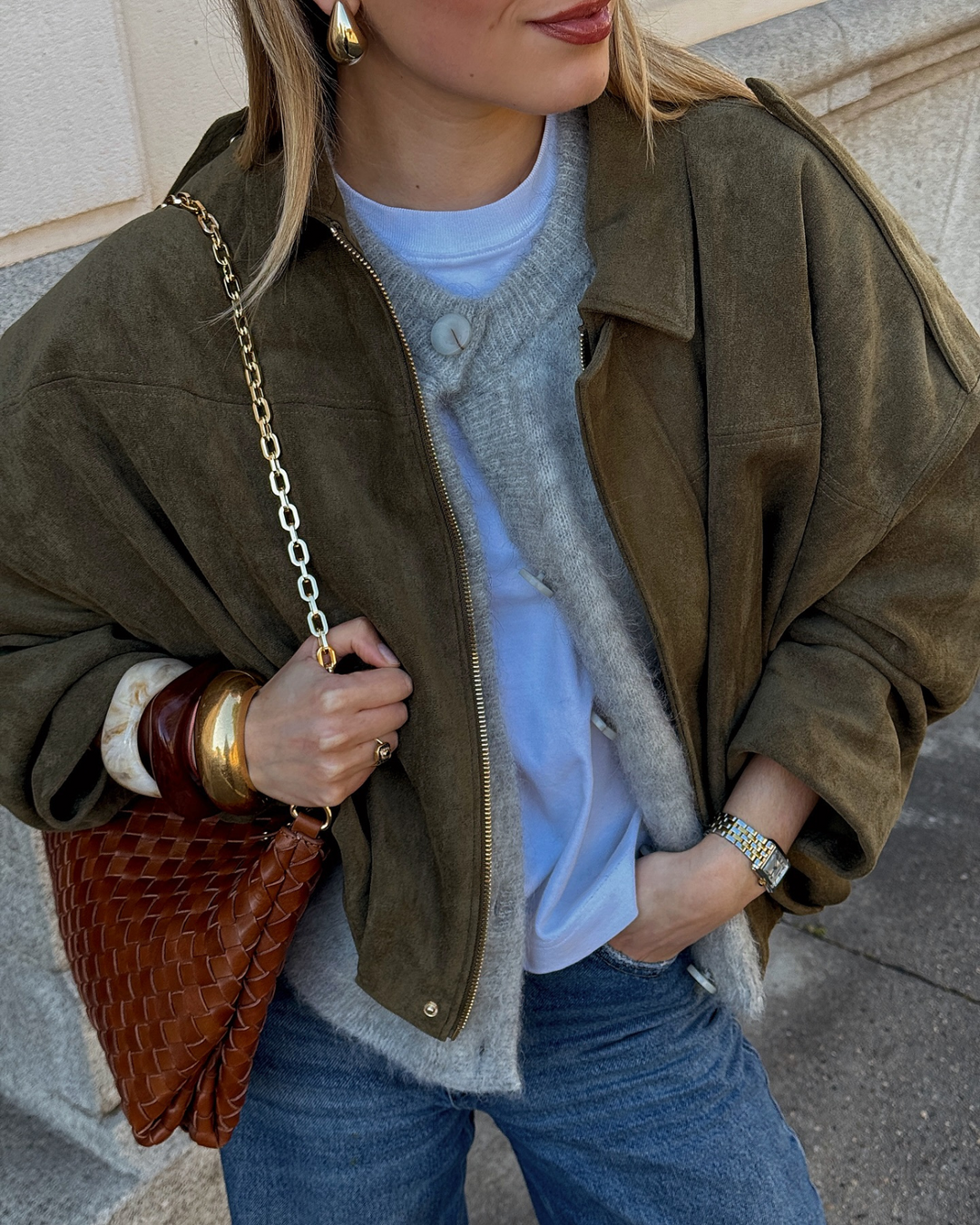 Woman wearing a green jacket, light blue sweater, and jeans, holding a brown leather bag.