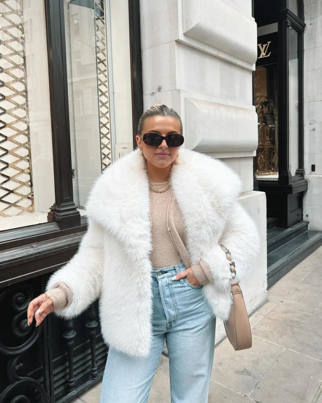 Person wearing a white fur coat, sunglasses, and light blue jeans in front of a building.