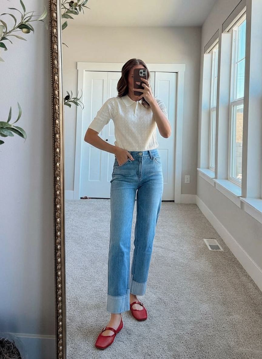 Person taking a mirror selfie wearing a white top and blue jeans in a room with large windows.