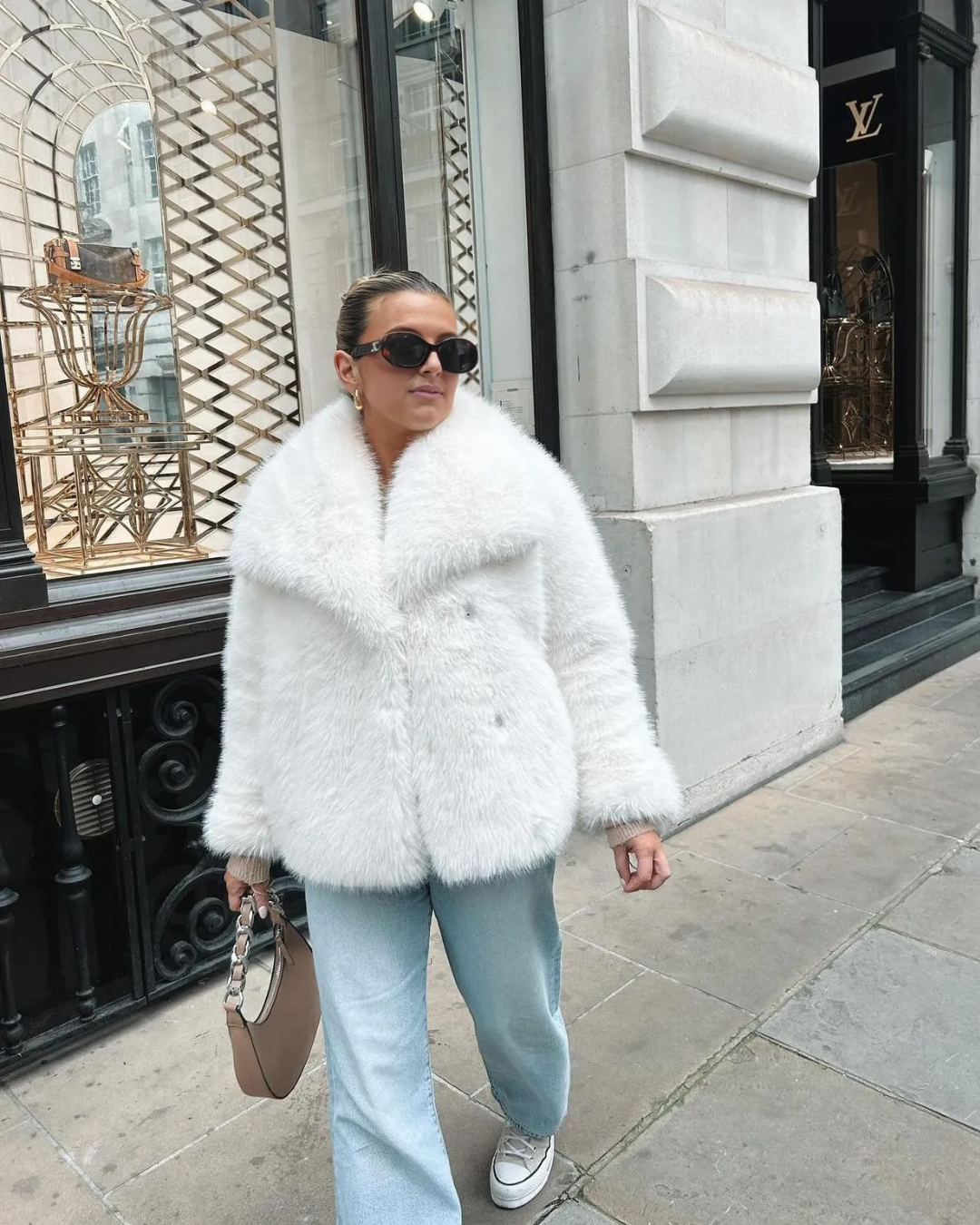 Person wearing a white fur coat and sunglasses on a city street.