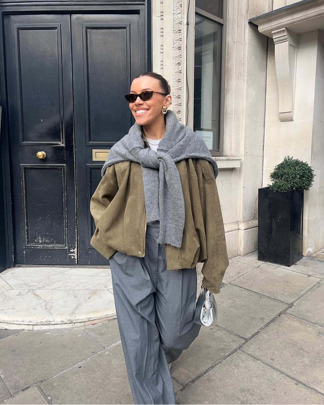 Woman walking on a sidewalk wearing a green jacket, gray scarf, and gray pants.