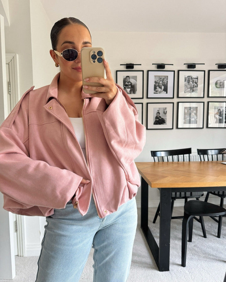 Person wearing a pink jacket and light blue jeans taking a mirror selfie in a room with a dining table and framed pictures on the wall.