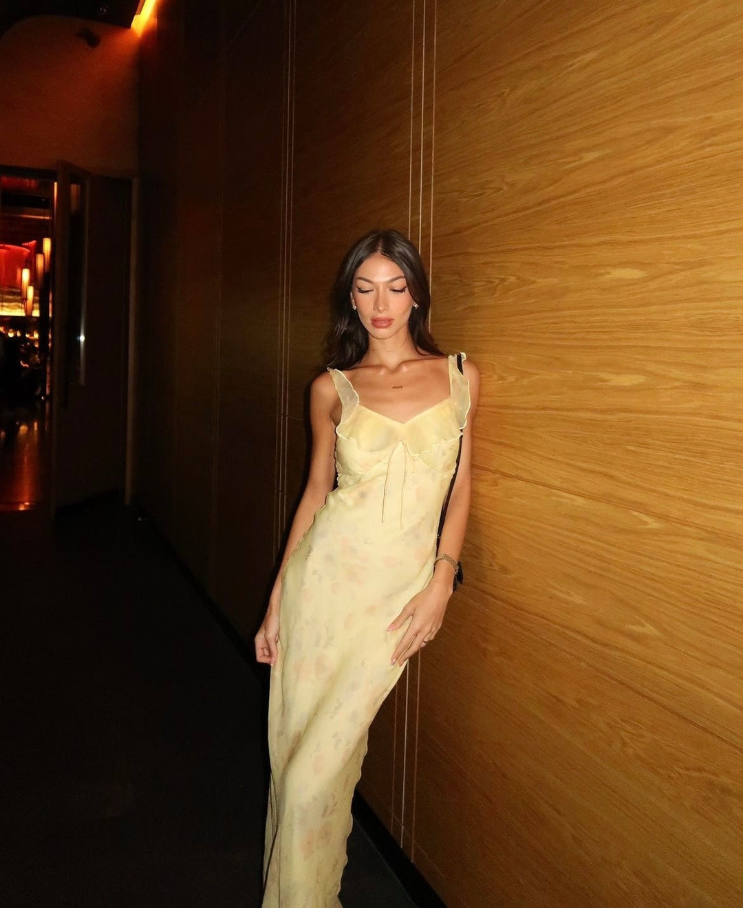 Woman in a light-colored dress standing against a wooden wall.