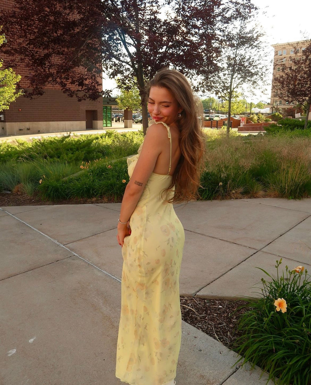 Woman in a yellow dress standing outdoors on a sidewalk with trees and grass in the background