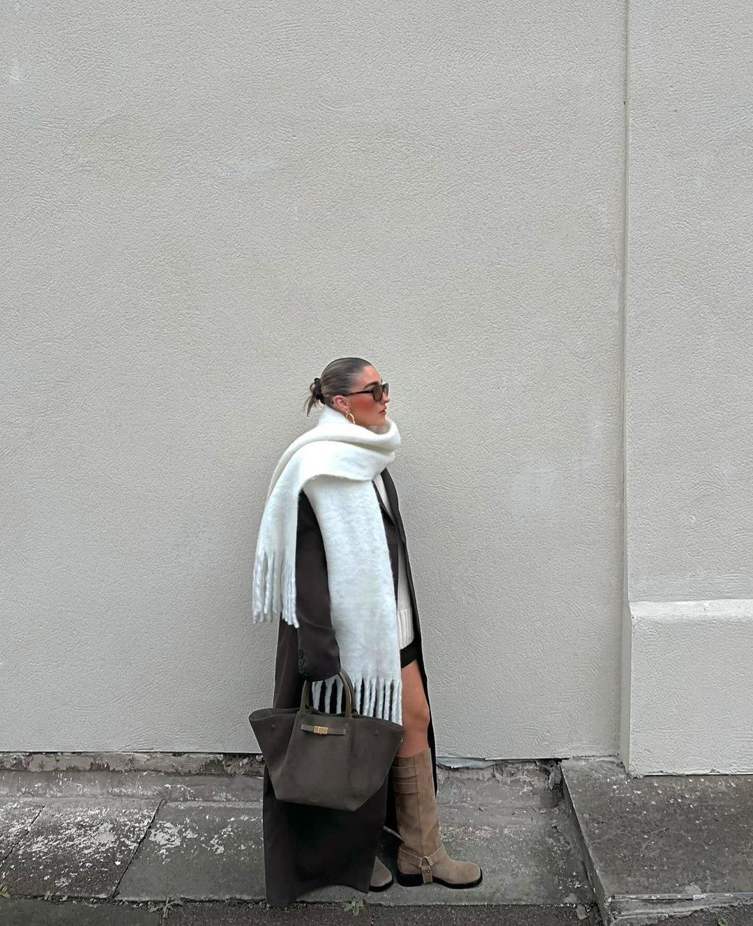 Person wearing a white scarf, dark coat, and brown boots against a plain wall.
