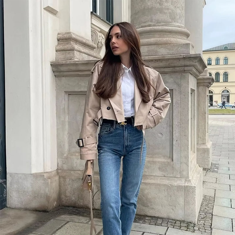 Woman in beige jacket and blue jeans standing in front of a classical building.