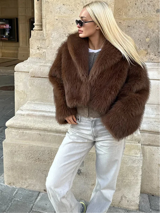 Person wearing a brown fur coat and white jeans standing against a stone wall.