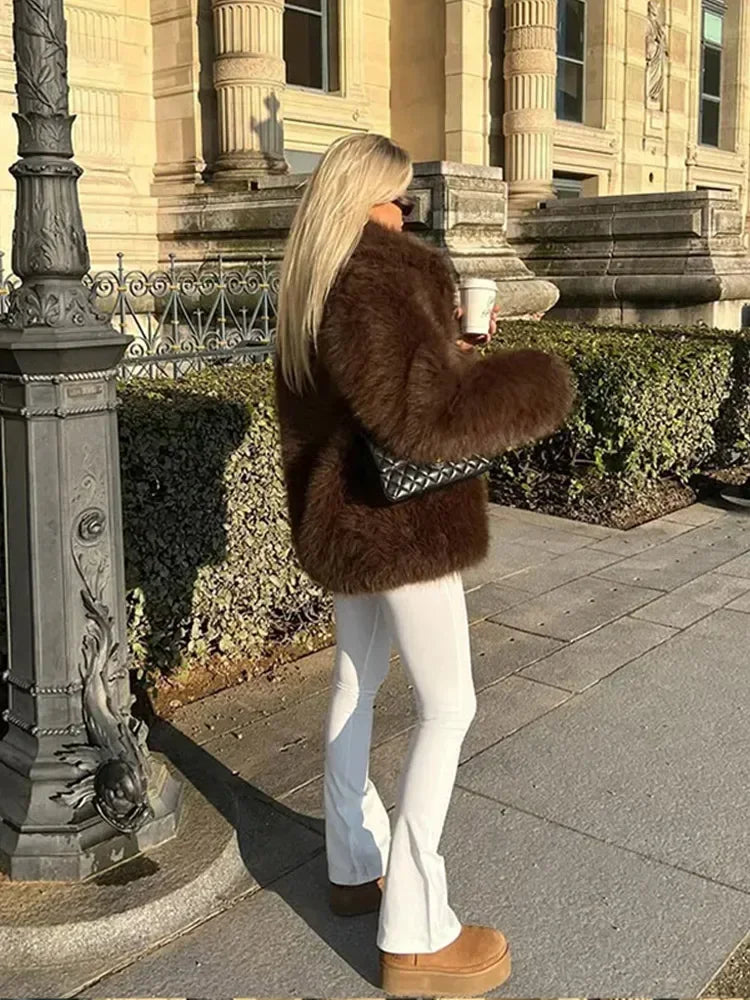 Person wearing a fur coat and white pants standing on a sidewalk with classical architecture in the background.