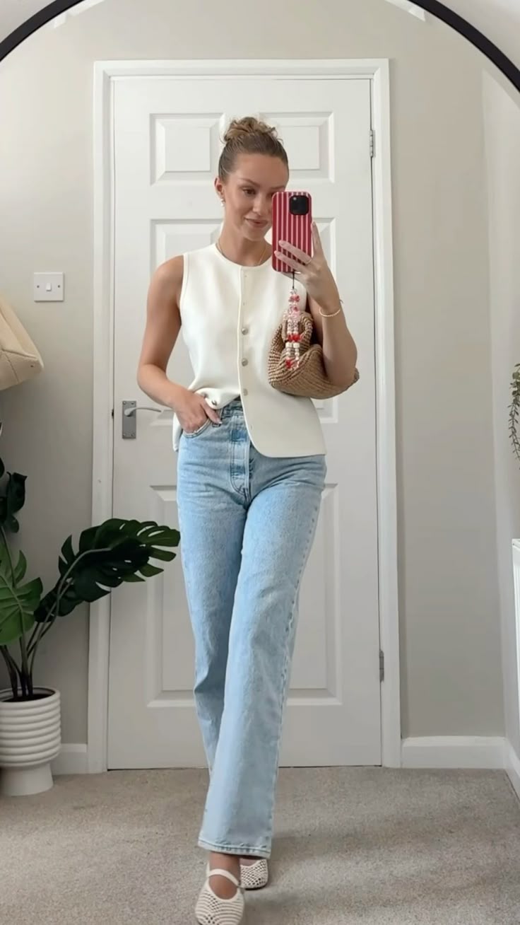 Woman taking a mirror selfie wearing a white sleeveless top and light blue jeans in a room with a door and plant.