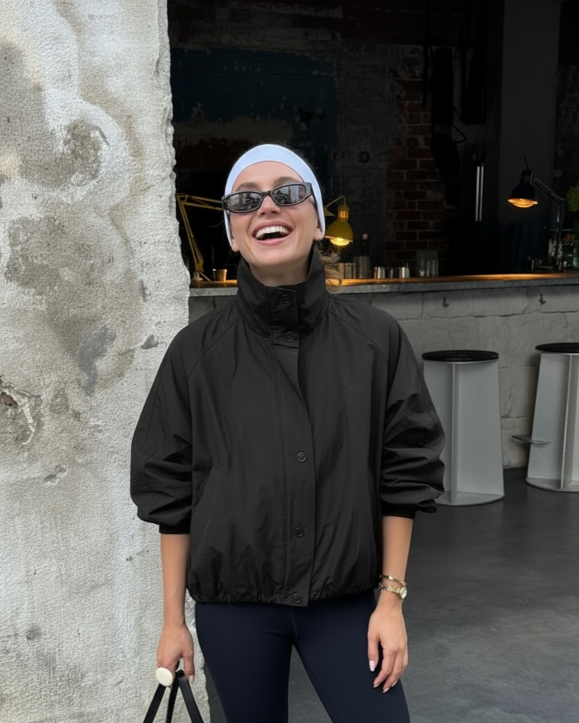 Person wearing a black jacket and sunglasses, standing against a textured wall with a modern interior background.