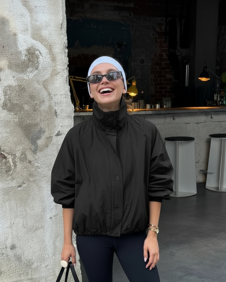 Person wearing a black jacket and sunglasses, standing against a textured wall with a modern interior background.