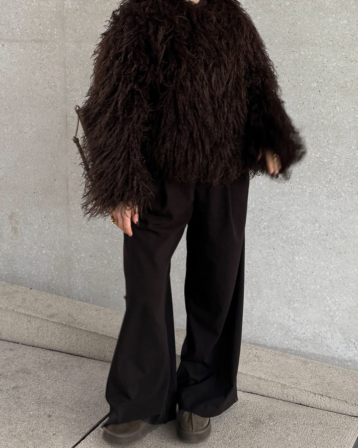 Person wearing a dark brown shaggy coat and black pants against a light gray wall.