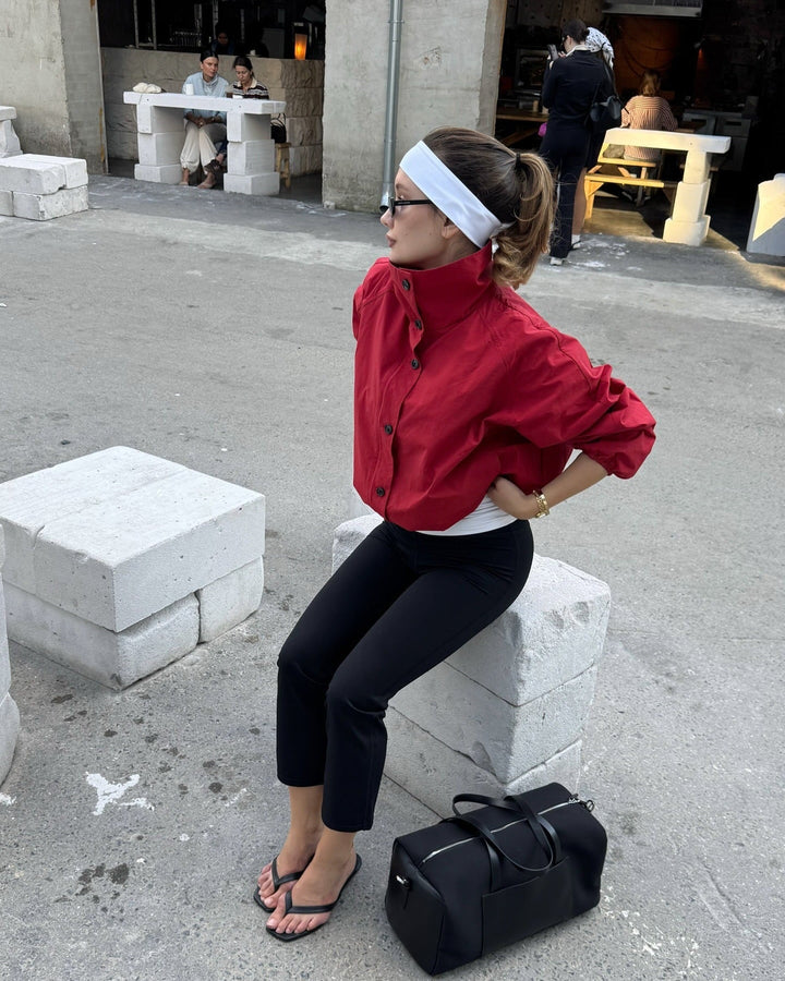 Woman in red jacket and black pants sitting on a concrete block with a black duffel bag nearby.