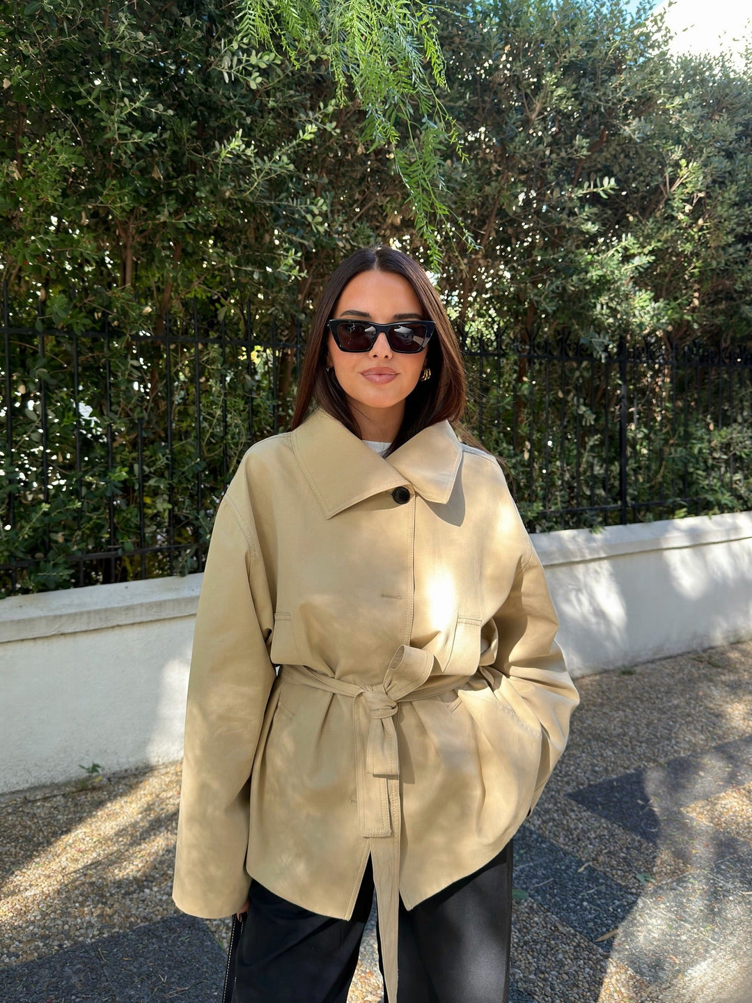 Woman wearing a beige trench coat and sunglasses outdoors with greenery in the background