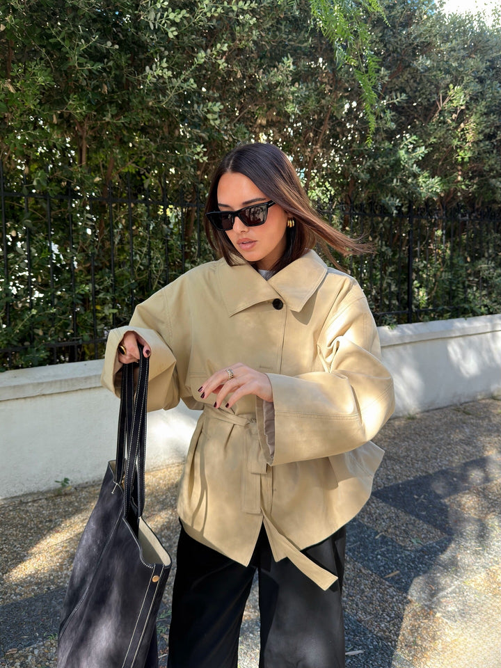 Woman in a beige coat holding a black bag outdoors with greenery in the background