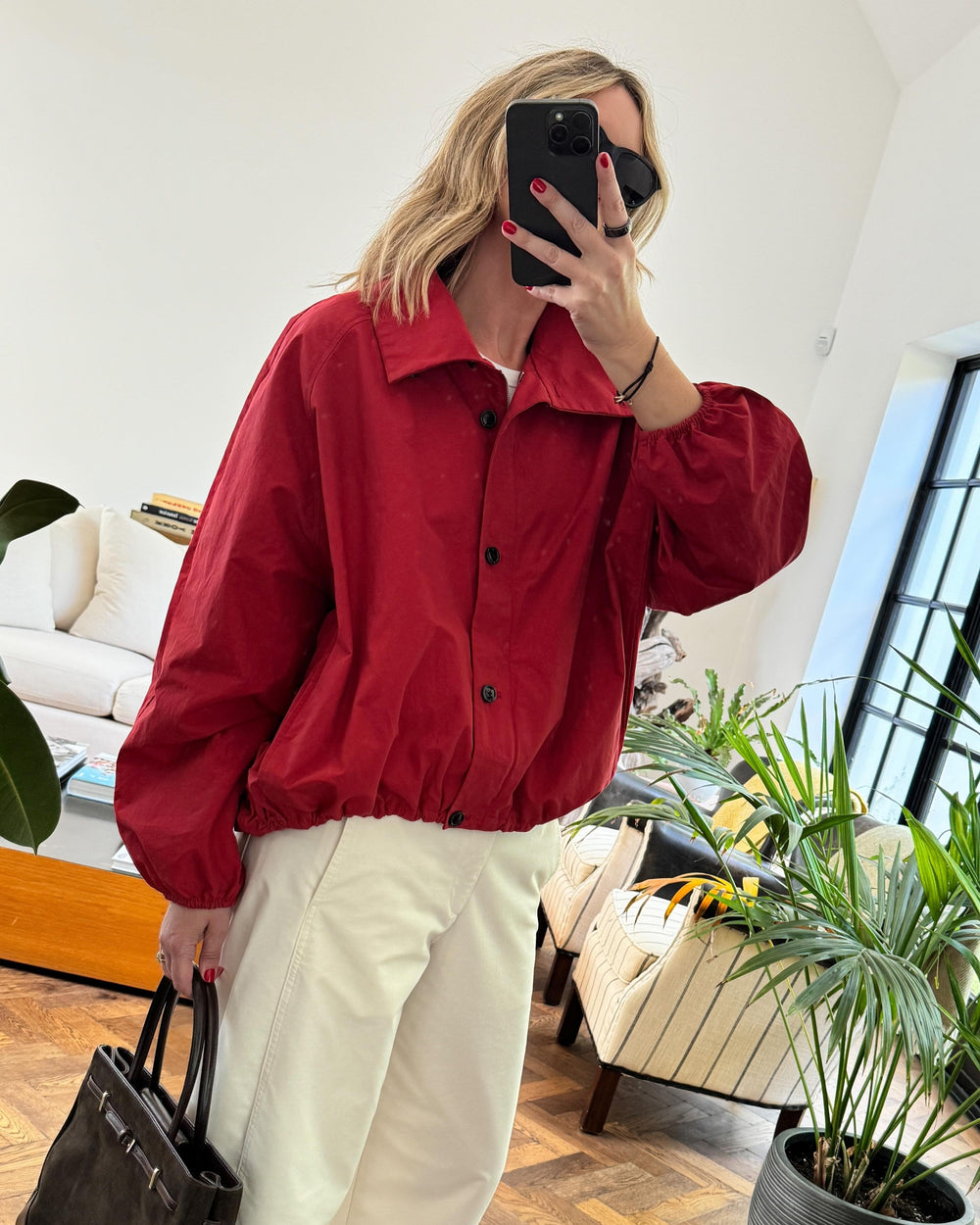 Person wearing a red jacket and white pants taking a mirror selfie in a living room.