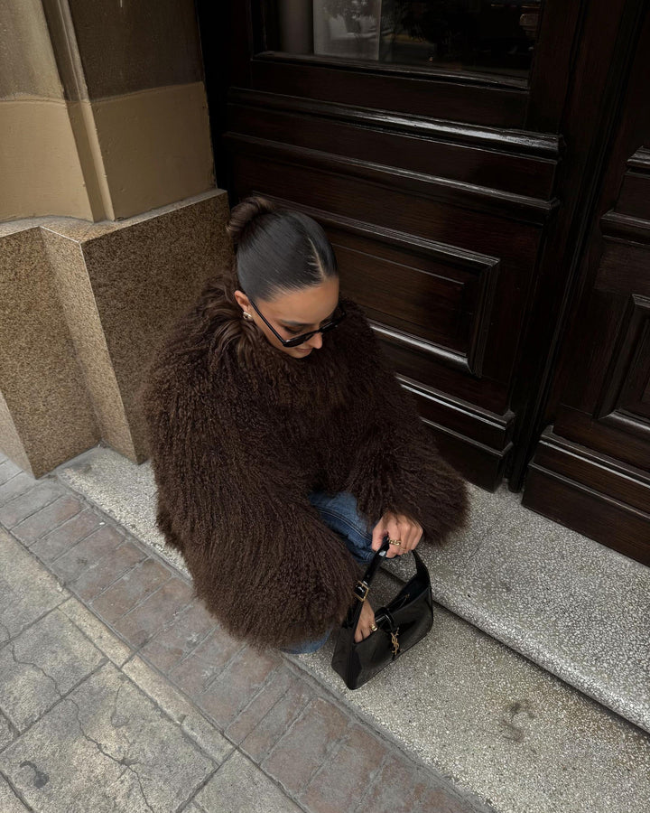 Woman in a brown fur coat sitting on a step with a black handbag.