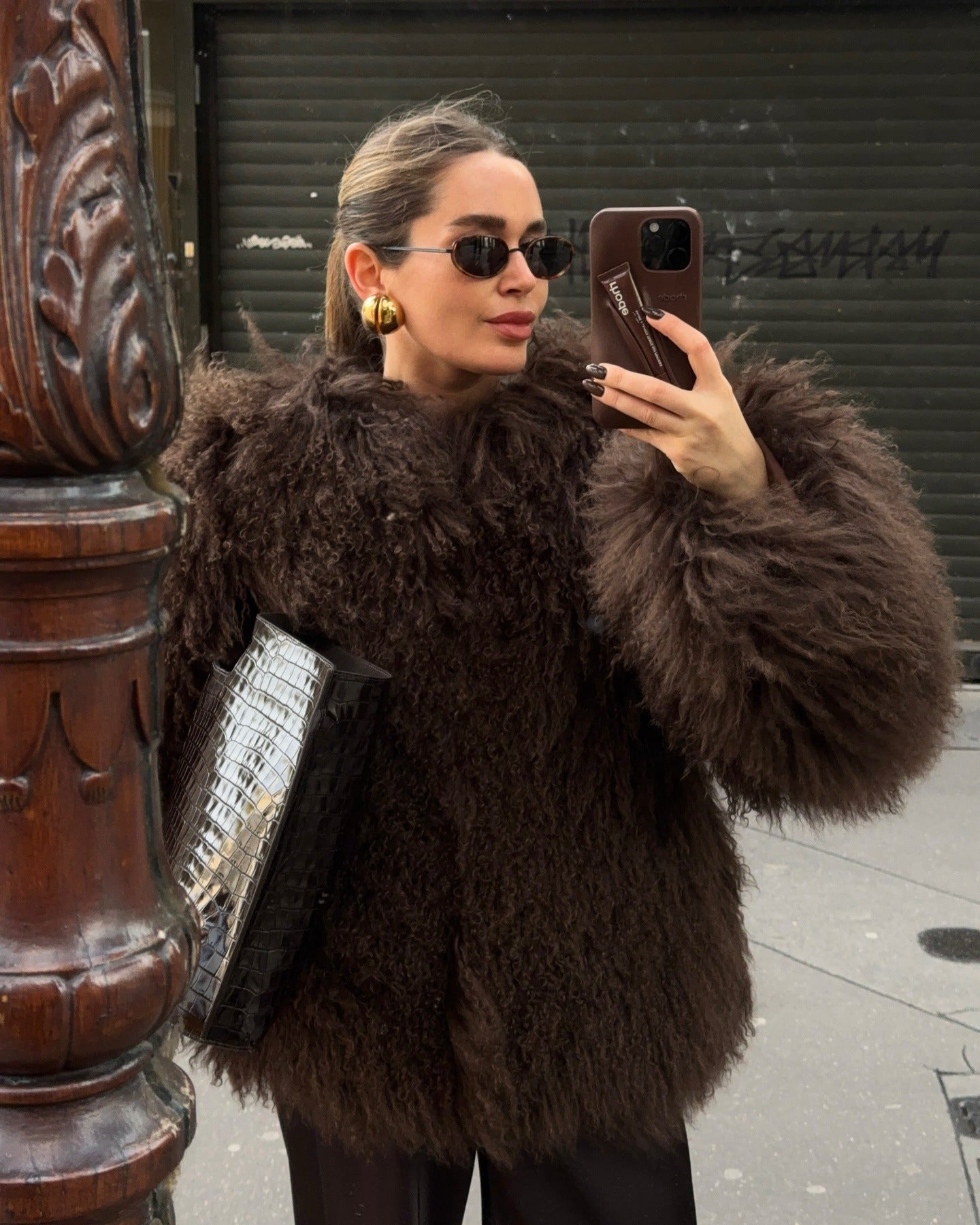 Woman taking a mirror selfie wearing a brown fur coat on a city street.