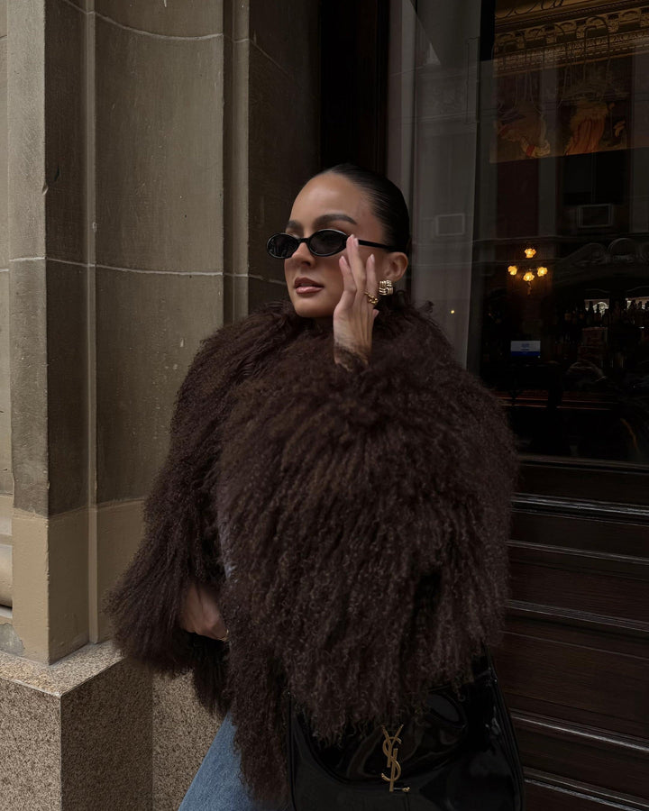 Woman wearing a dark brown fur coat and sunglasses, standing in front of a building entrance.